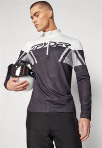 Zwart-witte sportieve longsleeve top met ritssluiting en kraag, voorzien van het "SPYDER" logo en hoekige designelementen, gecombineerd met een witte helm.