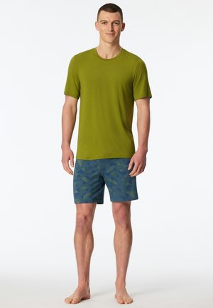 Man standing barefoot wearing a green short-sleeve t-shirt and blue patterned shorts against a plain white background.