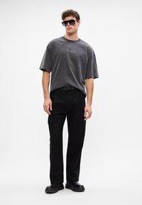 Graues, übergroßes T-Shirt mit "HUGO"-Logo, kombiniert mit schwarzen Cargo-Hosen mit Taschen und robusten schwarzen Schuhen. Das Model trägt dunkle Sonnenbrillen.