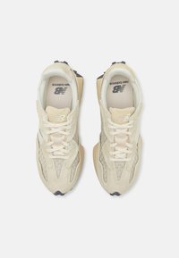 Ein Paar beiger New Balance Sneaker mit gewebter Textur, Wildlederüberzügen und gepolsterten Sohlen, von oben auf einem weißen Hintergrund betrachtet.