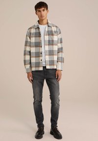 Camisa de franela en cuadros gris y crema con acentos marrones, que presenta botones y un bolsillo, llevada sobre una camiseta gris clara y unos vaqueros oscuros.