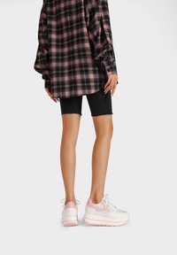 Sneakers bianchi e rosa con suola testurizzata, pannelli in rete e tallone imbottito. L'outfit include una camicia a quadri nera e pantaloni shorts in denim.