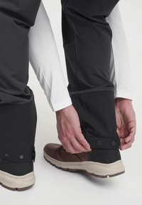 Pantalones de nieve negros con un diseño elegante, con un cierre de botón en el dobladillo, sobre zapatos marrones. Capas con una camisa de manga larga blanca.