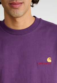 Camiseta de algodón morada con cuello redondo y logo bordado de "Carhartt" en amarillo y rojo en el área del lado izquierdo del pecho.