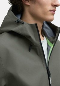 Chaqueta impermeable en verde oliva, con un alto collar, capucha ajustable, cremallera negra y un forro gris con un acento verde debajo.