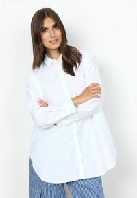 Camicia bianca a maniche lunghe con colletto a punta e vestibilità rilassata, abbinata a pantaloni a righe blu e bianche.