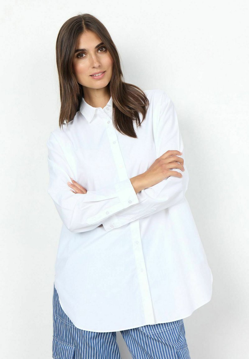 Camicia bianca a maniche lunghe con colletto a punta e vestibilità rilassata, abbinata a pantaloni a righe blu e bianche.