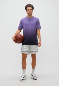 Νέος άνδρας με πουκάμισο σε μωβ όμπρε και λευκά σορτς μπάσκετ, κρατώντας μια μπάλα μπάσκετ, στέκεται μπροστά σε ένα απλό λευκό φόντο.