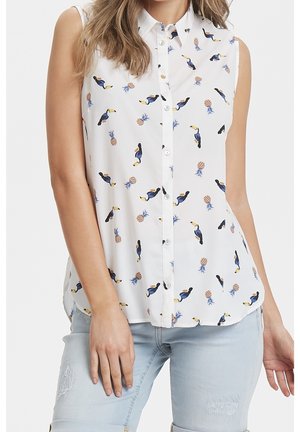 Blouse blanche sans manches boutonnée avec imprimé toucan et ananas, portée avec un jean bleu clair déchiré.