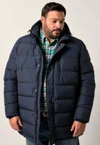 Hombre con una chaqueta de invierno acolchada azul marino sobre una camisa de cuadros verde y blanca, de pie contra un fondo claro y liso.