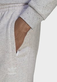 Hand i ljusgrå prickiga sweatpantsfickan med vitt Adidas-logotyp synlig nära fickan på höger ben.