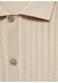 Camisa polo de punto en color beige con textura acanalada, que presenta un cuello clásico y un botón redondo gris en la tapeta frontal.