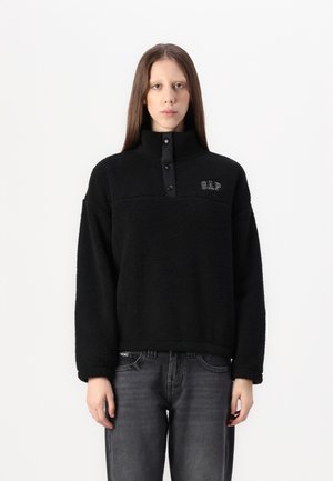 Pull en polaire noir avec col montant, fermeture à boutons-pression et logo GAP. Présente un tissu texturé et une coupe décontractée. Associé à un jean gris.