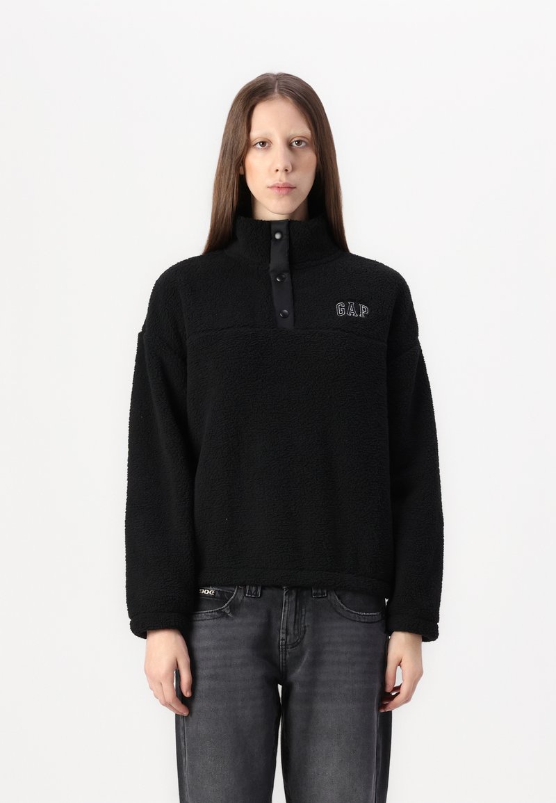 Zwarte fleece pullover met hoge kraag, drukknopen en GAP-logo. Kenmerkt zich door een gestructureerde stof en een relaxte pasvorm. Gecombineerd met grijze jeans.