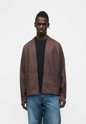 Fiatal férfi barna nyitott zsebes dzsekiben, fekete ing és kék farmer nadrág viselése mellett áll egy egyszerű világos háttér előtt.