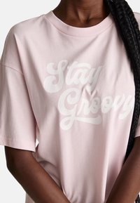 Svetle růžové bavlněné tričko s krátkým rukávem, na přední straně s bílým kurzivním textem "Stay Groovy". Hladká textura, uvolněný střih.