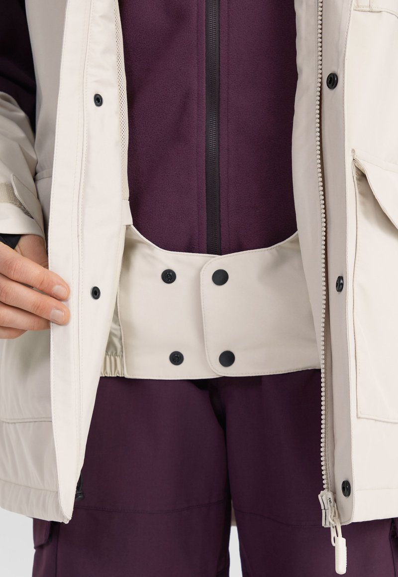 Beige Außenjacke mit einem Reißverschluss und schwarzen Druckknöpfen, über einer lila Fleece-Innenschicht. Enthält sichtbare Reißverschluss- und Taschen-details.