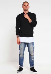 Camisa negra de manga larga, camiseta interior gris claro, vaqueros azules desgastados y zapatillas negras. El modelo mantiene una postura neutra.
