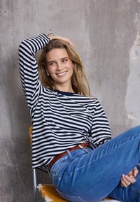 Merekollane ja valge triibuline pika varrukaga top ümmarguse kaelusega, siniste teksade ja pruuni vöökirjaga, istumas kollasel toolil vastakuti tekstuurse seina ees.