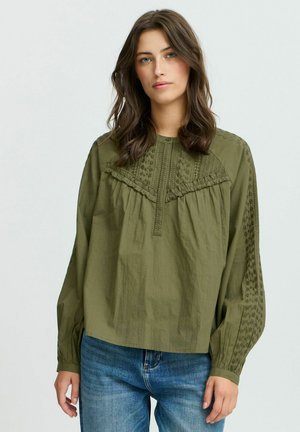 Blusa verde oliva con maniche lunghe, dettagli testurizzati, accenti a volant e una chiusura con bottoni; abbinata a jeans denim azzurri.