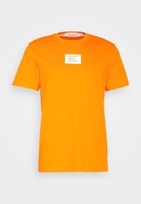 Camiseta de manga corta naranja brillante con un parche de logo rectangular blanco que dice "Calvin Klein Jeans New York 205 W 39 St." en el pecho.