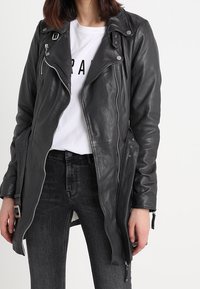 Veste de moto en cuir noir avec des zips argentés, des détails cloutés et un ourlet asymétrique. Associée à un t-shirt graphique blanc et un jean noir.