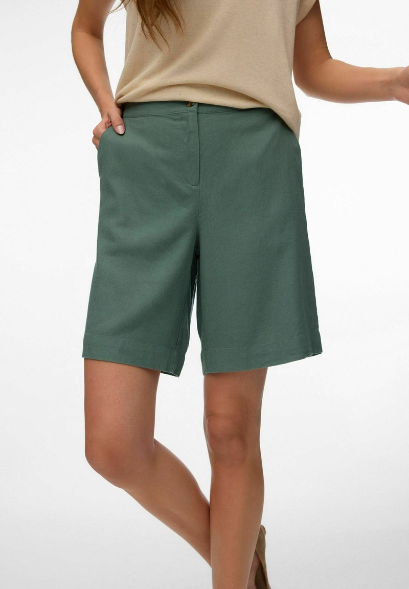 Femme portant un short vert jusqu'aux genoux et un haut beige, debout avec une main dans la poche, sur fond blanc.