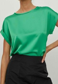 Blusa in satin verde con una vestibilità morbida, spalle basse e maniche arrotolate. Texture liscia con scollatura a girocollo. Abbinata a pantaloni neri.