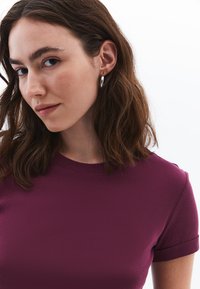 T-shirt bordeaux a maniche corte con scollo rotondo, caratterizzato da una texture liscia. Il modello ha capelli castani ondulati di media lunghezza e orecchini a cerchio in argento.