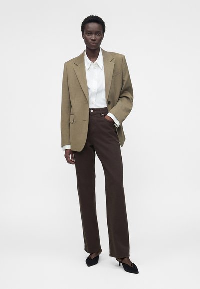 Μαύρη γυναίκα που φοράει μπεζ σακάκι, λευκό πουκάμισο με κουμπιά, σκούρο καφέ παντελόνι και μαύρα παπούτσια με τακούνι, στέκεται με το ένα χέρι στην τσέπη.