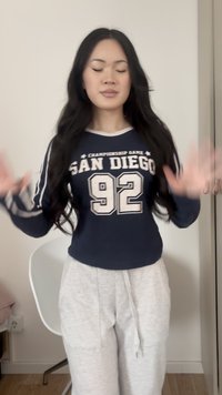 Woman with long black hair wears navy "San Diego 92" sweatshirt and gray sweatpants, standing indoors with eyes closed and hands raised.