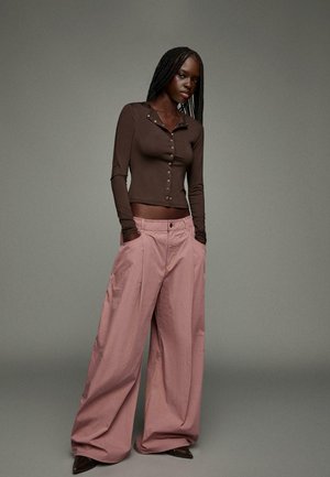 Braunes langärmliges Hemd mit Knöpfen, kombiniert mit übergroßen rosa Hosen. Die Hosen verfügen über seitliche Taschen und einen flachen Bund. Dunkle Absatzschuhe.