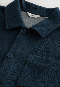 Camicetta blu navy con colletto, due bottoni neri e taschino sul petto. Il tessuto presenta un design a macchie sottili.