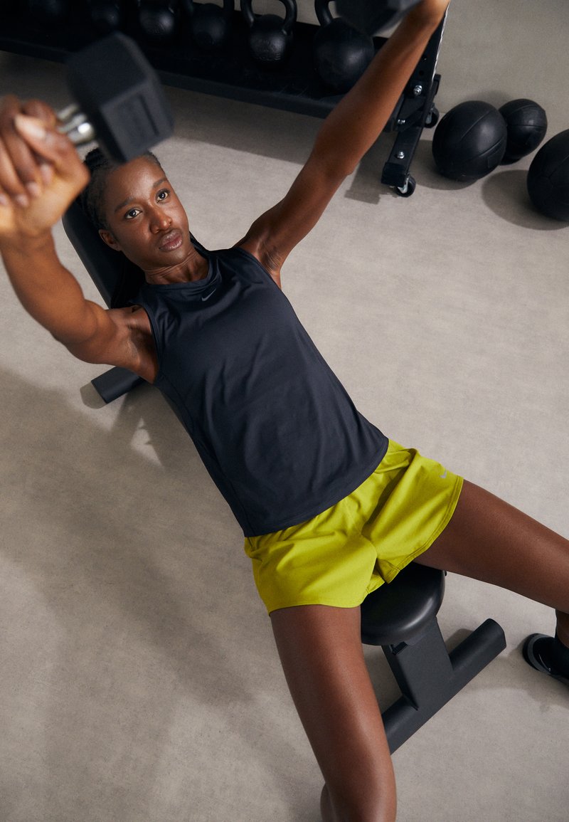 Schwarzes, ärmelloses Sportoberteil, neon-gelbe Shorts. Die Person hält eine Kurzhantel und sitzt auf einer Bank in einem Fitnessstudio, im Hintergrund sind Gewichte zu sehen.