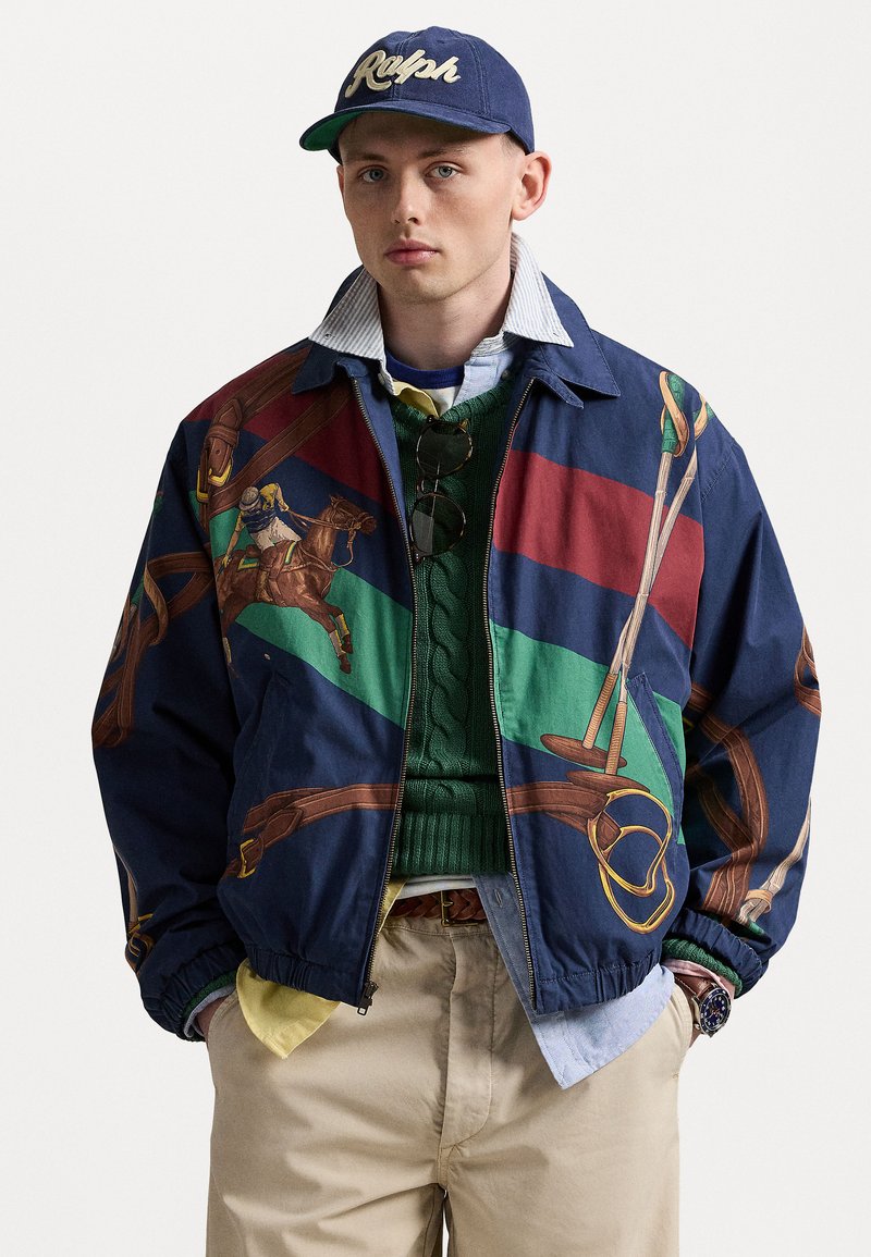 Coupe-vent marine avec des graphiques sur le thème de l'équitation, présentant des rayures vertes et rouges. Un pull en tricot vert en dessous, avec des chemises superposées visibles.