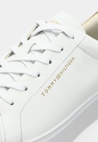 Baskets en cuir blanc avec un branding en or "TOMMY HILFIGER", présentant des lacets plats blancs et une texture lisse, un bout rond et un design minimaliste.