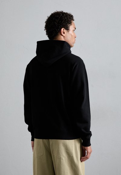 Sweat à capuche noir avec un cordon de serrage, manches et ourlet côtelés. Porté avec un pantalon beige clair, vu de dos sur un fond neutre.