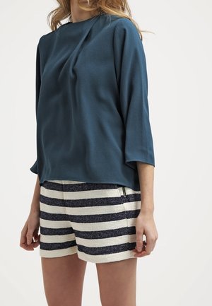 Blusa drappeggiata color teal con maniche a tre quarti abbinata a pantaloni corti a righe in bianco e navy. Texture notevole e design informale.