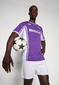 Maillot de football violet avec des accents et des logos blancs. Joueur tenant un ballon de football noir et blanc avec des motifs d'étoiles, portant un short blanc.