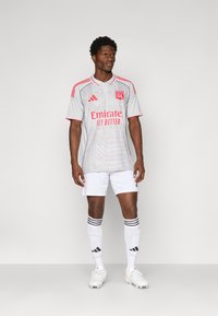 Athlète masculin portant un maillot de soccer gris de l'Olympique Lyonnais, un short blanc, des chaussettes blanches et des crampons blancs, se tenant devant un fond uni.