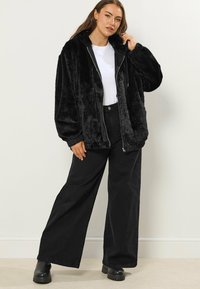 Czarna kurtka z futra sztucznego z zamkiem z przodu, kapturem i szerokimi rękawami, w połączeniu z czarnymi spodniami o szerokim kroju i masywnymi czarnymi butami.