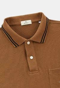 Polo shirt marron texturé avec un col côtelé et des rayures noires, une patte de boutonnage à deux boutons et une poche poitrine. L'étiquette indique "Serge Blanco."