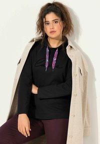 Felpa nera con cappuccio e coulisse, con accenti colorati, indossata sotto un cappotto beige chiaro. Il modello posa con un'espressione rilassata.