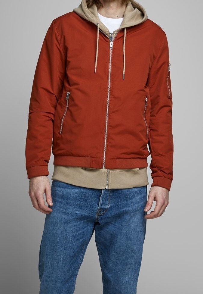 Homme portant une veste bomber rouge zippée sur un sweat à capuche beige et un jean bleu, debout devant un fond gris uni.