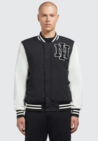 Zwart-witte varsityjas met een zwart nylon lichaam, witte mouwen, ribgebreide manchetten en een opvallend geborduurd logo op de borst.
