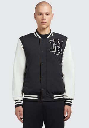 Zwart-witte varsityjas met een zwart nylon lichaam, witte mouwen, ribgebreide manchetten en een opvallend geborduurd logo op de borst.