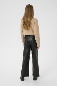 Woman with long hair wearing beige knit sweater, black high-waisted wide-leg leather pants, and black boots, standing against a white background.