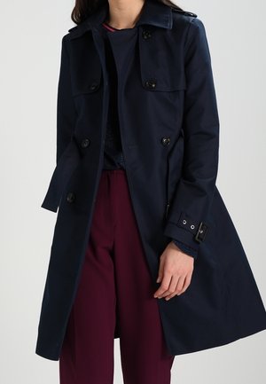 Kobieta ubrana w ciemnogranatowy trencz na czarnym topie i bordowych spodniach, stojąca na tle jednolitego jasnego tła.