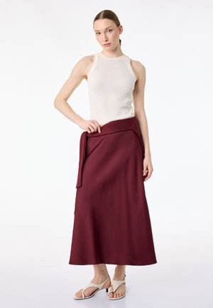 Kobieta stoi, ubrana w białą, bezrękawnikową prążkowaną bluzkę, bordową spódnicę midi z dopasowanym swetrem związanym w talii oraz beżowe sandały na otwartym palcu, na jednolitym białym tle.