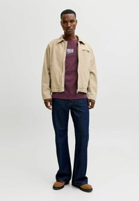 Beige bomberjakke over en vinrød skjorte, kombineret med mørkeblå vide jeans og brune støvler. Jakken har en frontlynlås og logodetaljer.